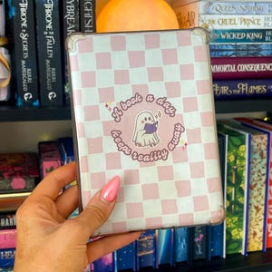 Op de afbeelding: Een doorzichtige beschermhoes voor een e-reader met een lichtroze en wit ruitpatroon. In het midden zit een ronde sticker met een witte geest die een paars boek leest, omringd door de tekst A book a day, keeps reality away.