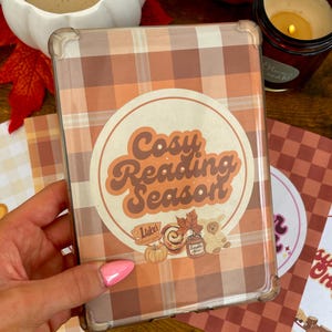 Könnte beinhalten: Eine rechteckige E-Reader-Hülle mit braun-cremefarbenem Karomuster. Ein runder Aufkleber in der Mitte zeigt den Text "Cosy Reading Season" in einer Retro-Schriftart, begleitet von kleinen Illustrationen eines Kürbisses, einer Zimtschnecke und eines Bären.