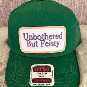 May include: A solid green trucker hat with a mesh back and a rectangular patch on the front. The patch has a white background with a light pink border and features the text Unbothered But Feisty in purple and pink lettering. An Otto brand sticker is attached.