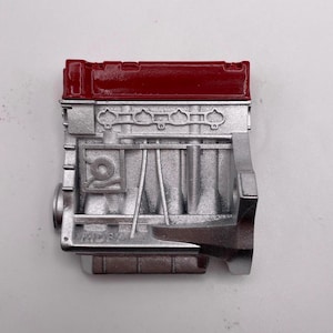 Op de afbeelding: Een gedetailleerd, zilverkleurig model van een motorblok met een glanzend rood bovenstuk. Het motorblok heeft ingewikkelde details, waaronder cilinders, kleppen en andere mechanische componenten. Het model is een miniatuurweergave van een automotor.