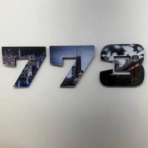 May include: Wooden wall art spelling out the number 773. Each number is a different piece, with a cityscape photograph filling the negative space. The numbers are mounted on a white wall.