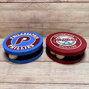 Può includere: Due espositori rotondi per palline da baseball dei Philadelphia Phillies. La custodia a sinistra è blu con un logo marrone che presenta una P bianca e il testo Philadelphia Phillies. La custodia a destra è marrone con un logo circolare e uno stadio.