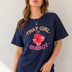 Peut inclure: Un t-shirt bleu marine à manches courtes avec le texte THAT GIRL en jaune, le nombre 777 en petite police jaune, un motif central de fleur d'hibiscus rouge et le mot ENERGY en rose, avec de petits accents en losange jaune sur le devant.