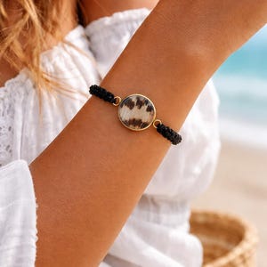 Pode incluir: Uma pulseira dourada com um pendente circular com um padrão de pele de vaca. O pendente está ligado a cordões trançados pretos. A pulseira é usada num braço, com um top branco visível no fundo. A praia é visível no fundo.