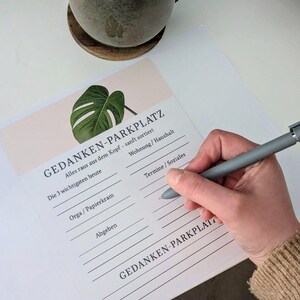 Könnte beinhalten: Eine Person schreibt mit einem grauen Stift auf einem Notizblock mit der Aufschrift "GEDANKEN-PARKPLATZ". Der Notizblock hat ein Monstera-Blatt-Design und fordert zur Organisation von Gedanken auf. Eine Keramiktasse und ein Untersetzer sind im Hintergrund.