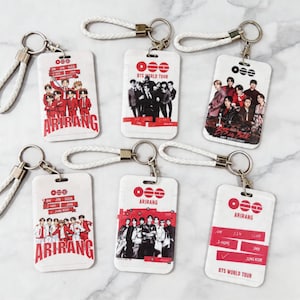 Puede incluir: Llaveros rectangulares blancos con varias imágenes de un grupo musical, con detalles en rojo y negro. Algunos llaveros tienen la palabra "ARIRANG" en rojo. Cada llavero tiene una correa de muñeca trenzada blanca y un cierre de metal plateado.