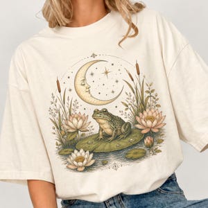 Könnte beinhalten: Ein cremefarbenes T-Shirt mit kurzem Arm und einem Grafikdruck, der einen Frosch auf einem Seerosenblatt zeigt, umgeben von Lotusblumen, Schilfkolben und einer Mondsichel mit Gesicht. Die Illustration verwendet gedeckte Erdtöne.