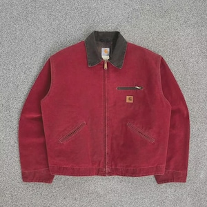 Op de afbeelding: Een rode jas met een donkerbruine kraag en een ritssluiting. De jas heeft twee voorzakken en een klein zakje op de borst. Een klein Carhartt-logo is zichtbaar op de borstzak.