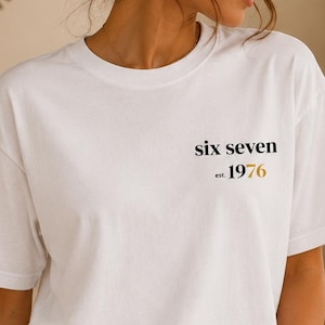 Könnte beinhalten: Weißes T-Shirt mit dem Text "six seven est. 1976" in Schwarz und Gold. Das Shirt hat kurze Ärmel und einen Rundhalsausschnitt. Die Person trägt ein goldenes Armband und Creolen.