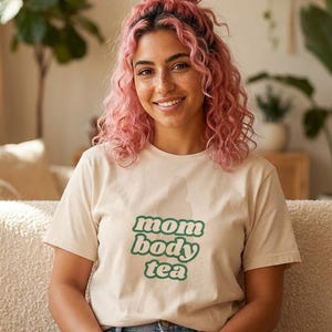 Peut inclure: Un t-shirt beige avec les mots "mom body tea" en vert, porté par une personne aux cheveux bouclés roses. La personne sourit et est assise sur un canapé beige dans une pièce bien éclairée avec des plantes.
