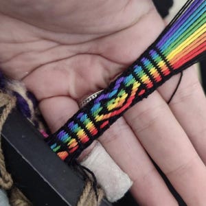 May include: A woven friendship bracelet featuring a vibrant rainbow-colored pattern against a black background. The design includes horizontal stripes and a central heart-shaped motif created with red, orange, yellow, green, blue, and purple thread.