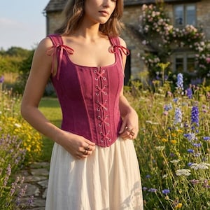 May include: A deep magenta corset-style top featuring a square neckline, vertical ribbed texture, and a front lace-up closure with silver-toned grommets. The garment includes thin ribbon shoulder straps that tie into bows at the top of each shoulder.