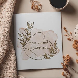 May include: A white square notebook with the text Mon carnet de grossesse printed in black. The cover features a watercolor-style beige heart shape outlined in black, decorated with green leafy branches on the left and right sides.
