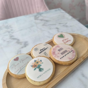 May include: A wooden tray holds six round cookies with white icing and various designs. The cookies feature floral bouquets, hearts, and text that reads "Merci Nounou". The cookies are arranged on a light-colored marble table.