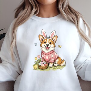 May include: White sweatshirt featuring a cartoon corgi wearing bunny ears and a pink sweater with Easter egg designs. The corgi is sitting next to a basket of Easter eggs and a baby chick. Butterflies and flowers are also in the image.