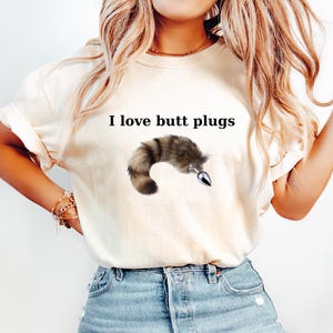 May include: Cream-colored t-shirt with the text "I love butt plugs" above a graphic of a furry tail with a silver plug. The shirt is paired with light blue denim jeans.