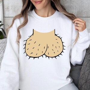 May include: White sweatshirt featuring a humorous graphic of a pair of testicles. The graphic is in a light tan color with black details and a hairy texture. The sweatshirt is a casual, unisex garment.