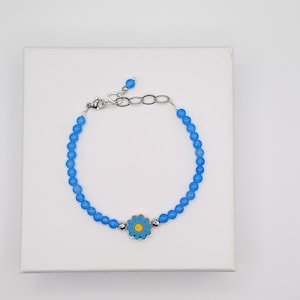 Pode incluir: Uma pulseira de contas azuis com fecho prateado e um berloque de margarida. O berloque de margarida tem um centro amarelo e pétalas azuis. A pulseira está sobre uma superfície branca.