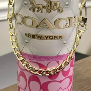Puede incluir: Un vaso blanco y rosa con un asa de cadena dorada. La sección superior presenta un diseño acolchado con el logotipo de Coach y las palabras "NEW YORK". La sección inferior tiene un patrón rosa.