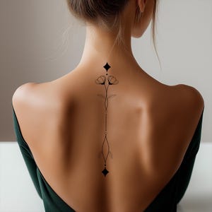 Può includere: Un disegno di tatuaggio floreale nero sulla schiena di una persona. Il tatuaggio presenta due fiori in alto, una linea di punti e una forma a diamante in basso. La persona indossa un top verde scuro.