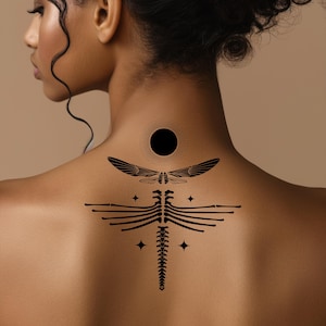 May include: Black temporary tattoo design on the back. The tattoo features a sun, dragonfly, rib cage, and stars. The design is symmetrical and detailed, with fine lines and shading. The tattoo is placed on the upper back and neck.