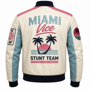 Könnte beinhalten: Cremefarbene Bomberjacke mit blauen und rosa Akzenten. Auf der Rückseite steht "Miami Vice" und "Stunt Team" mit einer Grafik von Sonnenuntergang und Palmen. Auf den Ärmeln befinden sich Aufnäher mit "The Fall Guy" und einem kreisförmigen Sonnenuntergangsdesign.