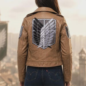 K&ouml;nnte beinhalten: Eine hellbraune Jacke mit einem gro&szlig;en gestickten Emblem auf dem R&uuml;cken, das zwei Fl&uuml;gel, einen schwarzen und einen wei&szlig;en, innerhalb einer Schildform zeigt. Kleinere Versionen desselben Fl&uuml;gel-Emblems sind auf beiden &Auml;rmeln aufgestickt.