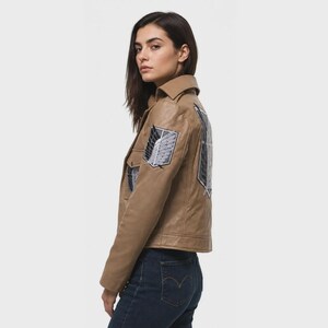 K&ouml;nnte beinhalten: Eine hellbraune Lederjacke mit spitzem Kragen, langen &Auml;rmeln und einer Knopfleiste. Die Jacke ist mit schwarz-wei&szlig;en Stickaufn&auml;hern auf der Brust und dem R&uuml;cken verziert, die ein schildf&ouml;rmiges Emblem mit Fl&uuml;geln und Schwertern darstellen.