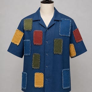 May include: A short-sleeved, blue button-down shirt with a camp collar. The shirt has rectangular patches in various colours, including yellow, green, red, and blue, with frayed edges. The shirt is displayed on a mannequin.