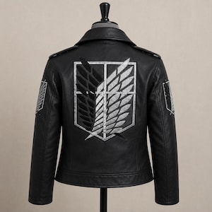 K&ouml;nnte beinhalten: Eine schwarze Lederjacke mit einem gro&szlig;en gestickten Emblem auf dem R&uuml;cken und kleineren passenden Emblemen auf jeder Schulter. Das zentrale Motiv zeigt einen Schild mit zwei &uuml;berlappenden Fl&uuml;geln, einem schwarzen und einem silbernen, sowie einer vertikalen Linie.