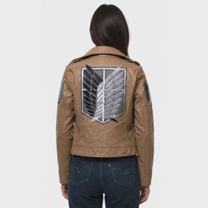 K&ouml;nnte beinhalten: Eine hellbraune Lederjacke mit einem gro&szlig;en gestickten Emblem auf der R&uuml;ckseite. Das Emblem zeigt zwei stilisierte Fl&uuml;gel, einen schwarzen und einen wei&szlig;en, in einem schildartigen Wappen. Die Jacke hat lange &Auml;rmel und einen Kragen.