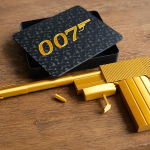 Op de afbeelding: Een goudkleurig speelgoedpistool samengesteld uit een pen, een aansteker en een manchetknop, vergezeld van een gouden kogel. Achter het pistool ligt een zwart rechthoekig blikken deksel met een geometrisch patroon en de gouden tekst 007.