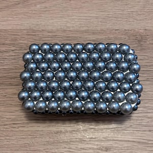 May include: A rectangular, beaded clutch featuring numerous gray-colored, spherical beads. The beads are tightly arranged, covering the entire surface of the clutch. The background is a light brown wooden surface.