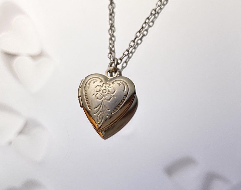Pu&ograve; includere: Una collana con un ciondolo a forma di cuore argentato con un design floreale. Il ciondolo ha un'apertura a cerniera e una catena delicata.