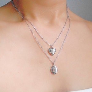 Pu&ograve; includere: Due collane d'argento con un ciondolo a forma di cuore e un ciondolo ovale. Entrambi i ciondoli presentano disegni intricati.