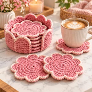 May include: A set of round, flower-shaped crochet coasters in a dusty rose pink color with a cream-colored border. The coasters feature a circular, textured stitch pattern and are stacked inside a matching crochet holder, with two coasters placed flat.