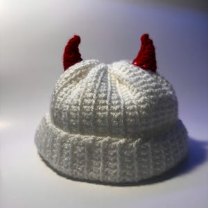 Puede incluir: Un gorro blanco tejido con dos cuernos rojos. El gorro está hecho de un hilo grueso y texturizado y tiene un borde doblado. Los cuernos rojos pequeños y puntiagudos añaden un toque juguetón al gorro.