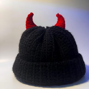 Puede incluir: Un gorro negro tejido con dos cuernos rojos. Los cuernos están curvados y sujetos a la parte superior del gorro. El gorro está hecho de un hilo grueso y texturizado. El gorro es de color oscuro y probablemente hecho a mano.