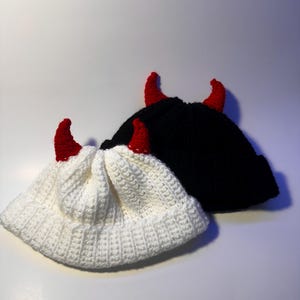 Puede incluir: Dos gorros de ganchillo, uno blanco y otro negro, cada uno adornado con cuernos de diablo rojos. Los sombreros están hechos de un tejido de punto acanalado texturizado. Los cuernos son de un rojo vibrante, contrastando con los colores de los sombreros.
