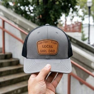 May include: Gray and black trucker hat with a curved brim. The hat features a leather patch with the text "SUPPORT YOUR LOCAL GIRL DAD". The hat is held by a person.