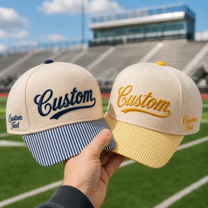 May include: Two baseball caps with cream-colored crowns and embroidered text. The left cap features navy blue text reading Custom and a navy blue striped brim. The right cap features yellow text reading Custom and a yellow striped brim.