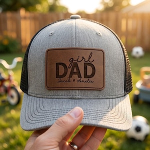 May include: A gray trucker hat with a black mesh back and a brown leather patch. The patch reads "girl DAD" with the names "Jacob & Amelia" below. The hat is held in a hand.