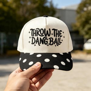 May include: A cream-colored baseball cap featuring black embroidered text that reads THROW THE DANG BALL surrounded by small star accents. The cap has a black brim decorated with a white polka dot pattern across the entire surface.