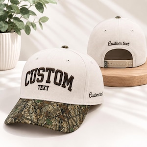 May include: A cream-colored baseball cap with a camouflage print brim. The front features the dark brown embroidered text "CUSTOM TEXT" in an arched font. The side displays "Custom text" in script, and the back has "Custom text" embroidered.