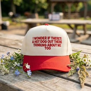 May include: A cream-colored trucker hat with a red brim. The hat features the embroidered phrase, "I WONDER IF THERE'S A HOT DOG OUT THERE THINKING ABOUT ME TOO" in red. The hat is resting on a wooden surface with flowers.