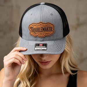 Pode incluir: Um bon&eacute; trucker cinza mesclado com tela preta na parte de tr&aacute;s e um patch de couro marrom claro na frente. O patch apresenta uma borda ornamentada e a palavra TROUBLEMAKER em preto. Um adesivo preto e branco na aba diz RICHARDSON 112.