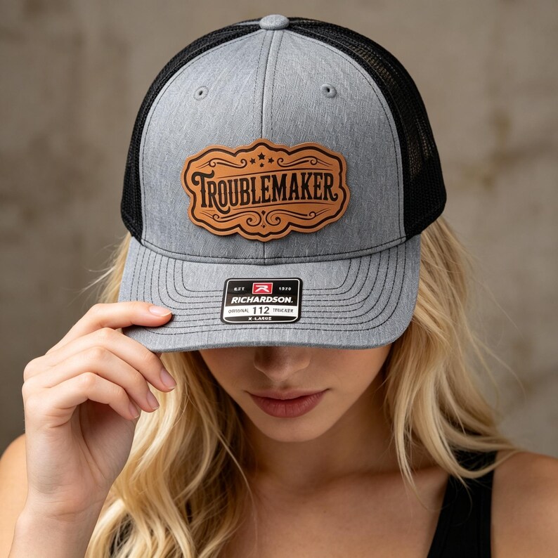 Pode incluir: Um bon&eacute; trucker cinza mesclado com tela preta na parte de tr&aacute;s e um patch de couro marrom claro na frente. O patch apresenta uma borda ornamentada e a palavra TROUBLEMAKER em preto. Um adesivo preto e branco na aba diz RICHARDSON 112.