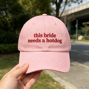 May include: A light pink baseball cap featuring the embroidered red text "this bride needs a hotdog" centered on the front panel. The cap has a structured crown, a curved brim, and matching pink stitching details across the fabric surface.