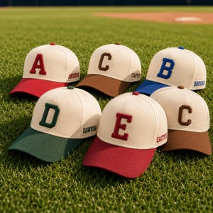 May include: Six baseball caps with cream-colored crowns and contrasting colored brims. Each cap features a large, raised letter on the front and a name embroidered on the side. The colors include red, brown, blue, dark green, and maroon accents.