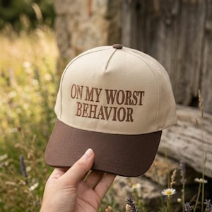 May include: A beige and brown baseball cap with the embroidered text "ON MY WORST BEHAVIOR". The cap has a curved brim and a button on top. The cap is held up by a hand.